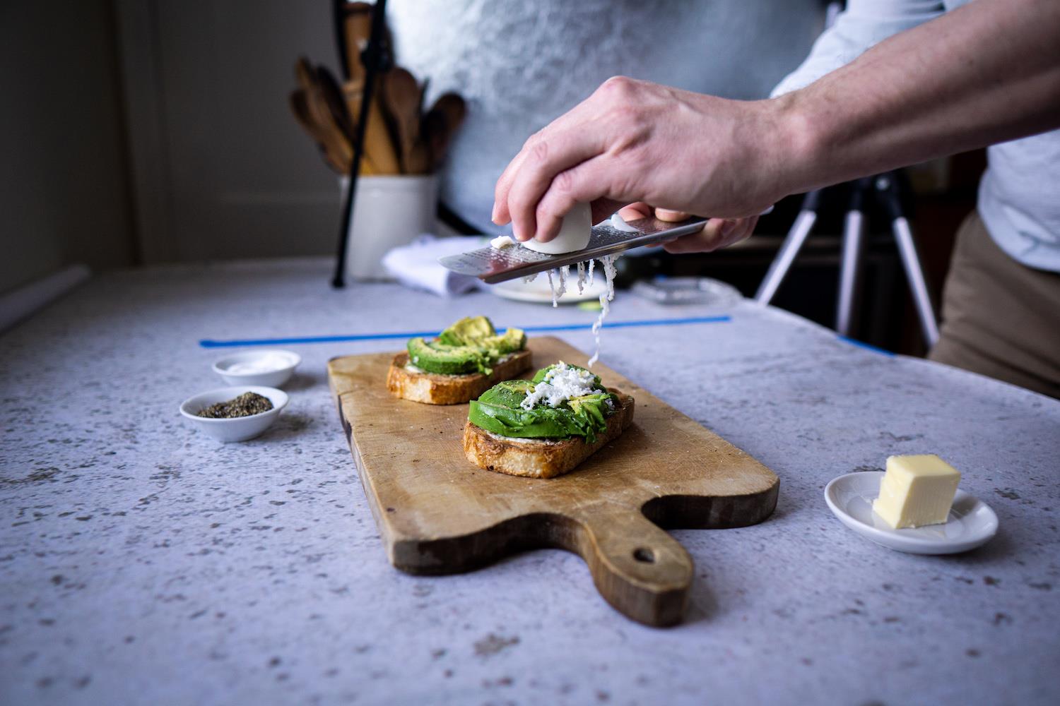 A pair of hands shaves a hardboiled egg onto avocado toast.